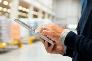 A person holding and using a tablet in a bright, industrial warehouse environment, focusing on digital operations or supply chain management